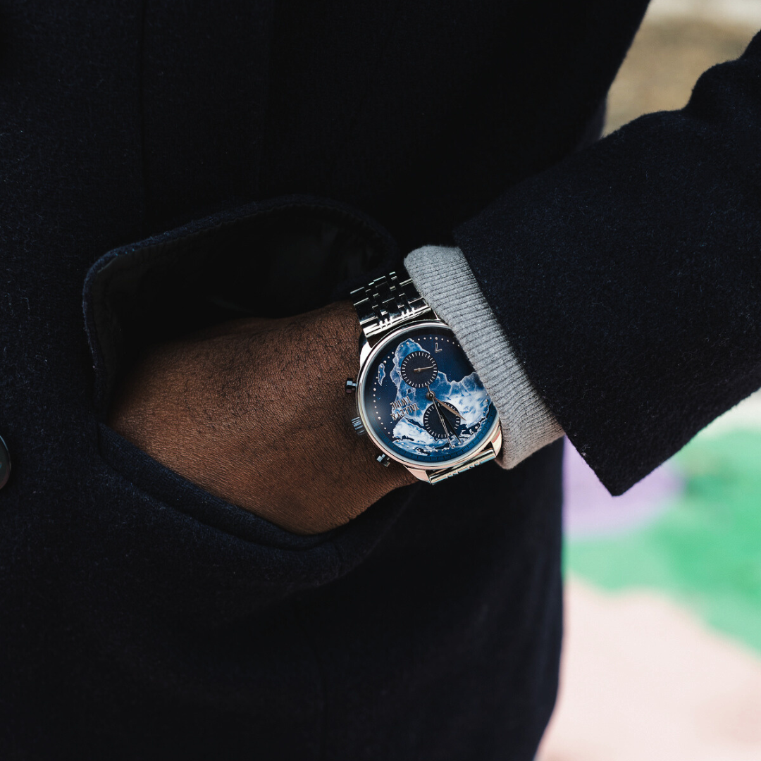 Origins watch with a blue dial featuring a moon phase display, worn on a wrist with a black coat sleeve.