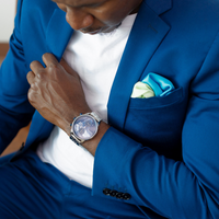 Silver watch with a blue dial worn on a man's wrist, paired with a blue suit and pocket square.